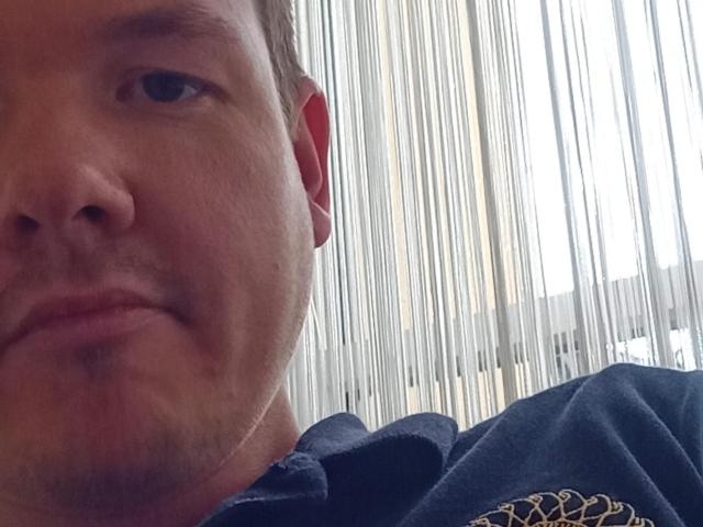 O rosto de ColinFunny, mostrando uma expressão neutra com os lábios ligeiramente entreabertos. Ele está vestindo uma camisa azul escura com bordados dourados. Sua pele é clara e tem pelos faciais leves..