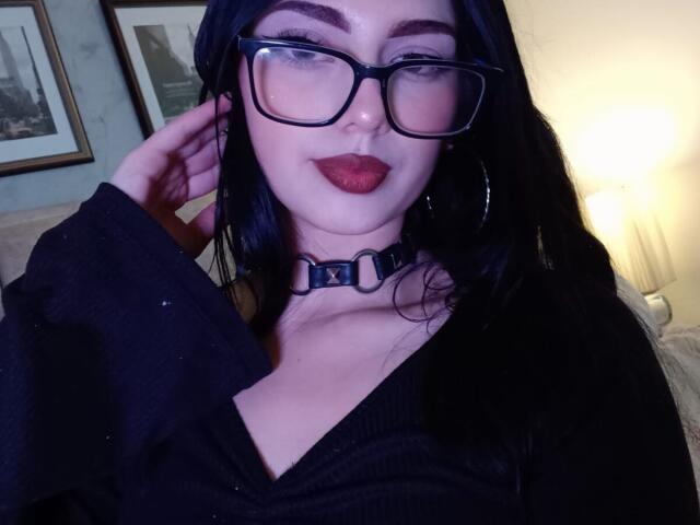 Isabella Rossi met zwart haar, een bril, rode lippenstift, een zwarte choker en een zwarte top, die haar borsten laat zien, met haar hand in haar haar, in een zacht verlichte kamer..