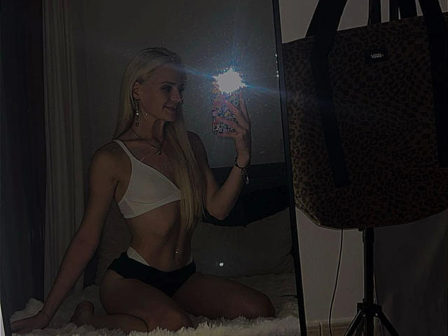 Mirror selfie of katanakiki-hot with long blonde hair. She's kneeling on a white, fluffy rug, wearing a white bralette and black lace panties. Her right hand holds a phone with a bright flash, reflecting in the mirror. A leopard-print bag is on a stand to the right. katanakiki-hot's slim, toned physique is highlighted by the lighting. The phone case has a colorful, patterned design. The image has a dark, low-light ambiance.