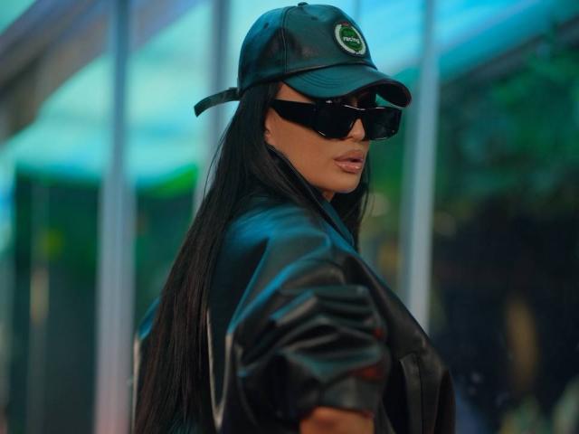 AstridVex with black leather jacket, open to show heart-shaped pasties covering her tits, black cap, smiling, arms crossed, standing in front of blurred green and blue background.