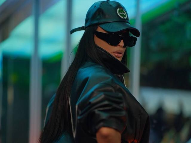 AstridVex in a black leather cap and sunglasses, pulling up a zipped jacket to reveal bare tits, with a green emblem on the cap. Tongue slightly out.