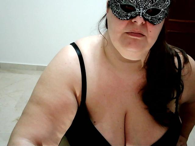 A photograph of PassionD. She's wearing a black lace mask with intricate patterns and a black spaghetti strap top, revealing ample cleavage. Her dark hair is partially visible, and she has a slightly flushed face. The image is focused on her upper body, with a beige tiled floor in the background. Her skin is light with some redness. The composition is close-up and slightly tilted.