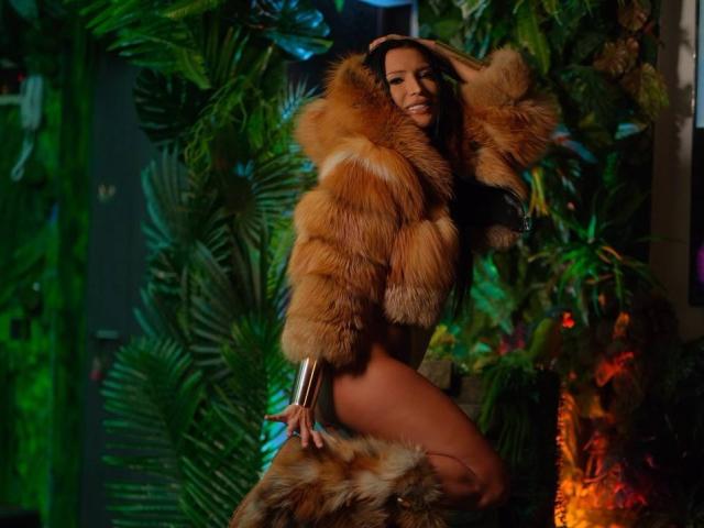AstridVex in a furry orange hood, long black hair, holding metal cuffs, wearing a gold mesh thong, standing with hands on hips, surrounded by green tropical leaves.