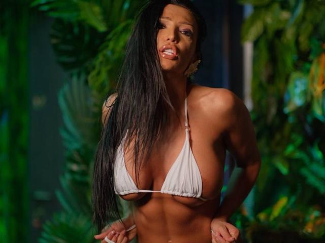 AstridVex with long black hair, wearing a white string bikini, showcasing her big tits and toned abs, standing in a lush green jungle. Her mouth is slightly open.