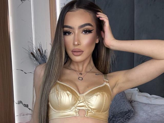 ClaraBenatti, con lunghi capelli castani, reggiseno dorato e trucco, mostra il seno, con una mano tra i capelli, mentre guarda la fotocamera..