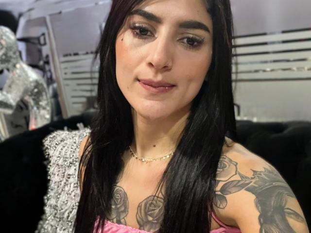 Ashley Scotts a les cheveux noirs, les yeux marrons et des tatouages sur les épaules. Elle porte un haut rose et un collier en or. Ses seins sont petits et elle sourit légèrement..