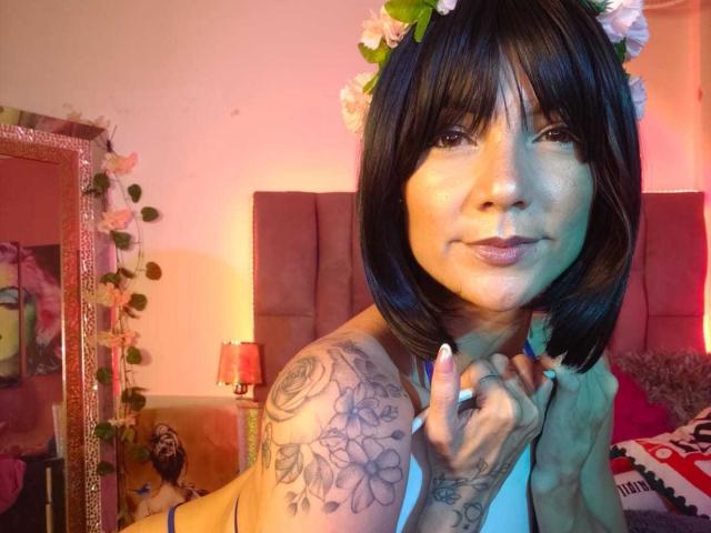 SweetYose has black bob hair, wears a flower headband, and shows off tattooed tits with a cigarette in a pink room. Her ass is partially visible on a bed.