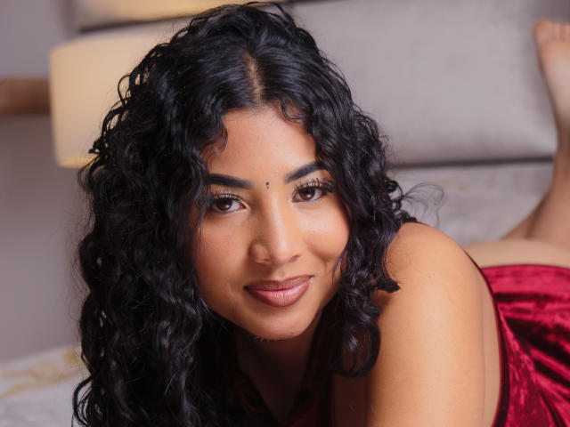 AntonelaRios has curly black hair, brown eyes, and a small beauty mark above her right eyebrow. She's lying on a bed in a red velvet top, smiling.