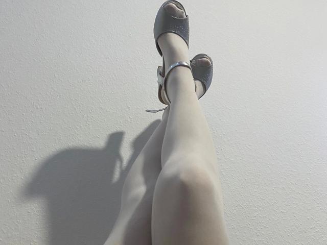 Close-up of RedObsession-hot's legs crossed at the ankles. She's wearing glittery, black, open-toe heels with a small strap. Her skin is pale with a slight shadow on the left. The heels cast a shadow on the white wall. The image is sharp with a focus on the legs and shoes, showing texture on the wall and smoothness of the legs. The composition is simple and direct.