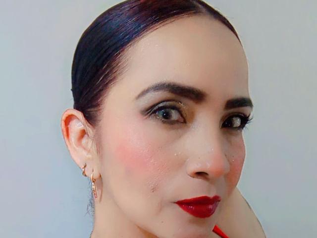 IsabellaMillerXx has shiny brown hair, red lipstick, and subtle makeup with pink blush. She wears gold earrings and has a slight smile, with visible freckles on her nose.