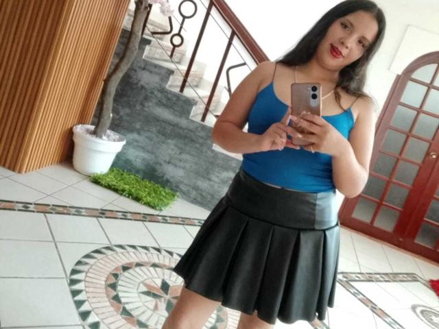 RosieTurner with long black hair, blue top, black pleated skirt, holding phone, smiling, red lipstick, medium tits, standing in tile room, wooden wall, potted plant, red door.