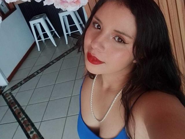RosieTurner with long dark hair, red lipstick, blue top, showing small tits, necklace, and a cute smile, standing on tiled floor, stools in background, selfie angle.