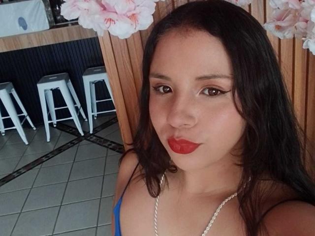 RosieTurner with dark hair, red lipstick, and a blue top, showing off her tits, standing near white bar stools and pink flowers in a tiled room.