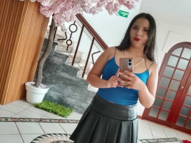 RosieTurner with dark hair, red lipstick, blue tank top, and black pleated skirt, holding a phone, standing in front of a cherry tree. Her tits are medium-sized.