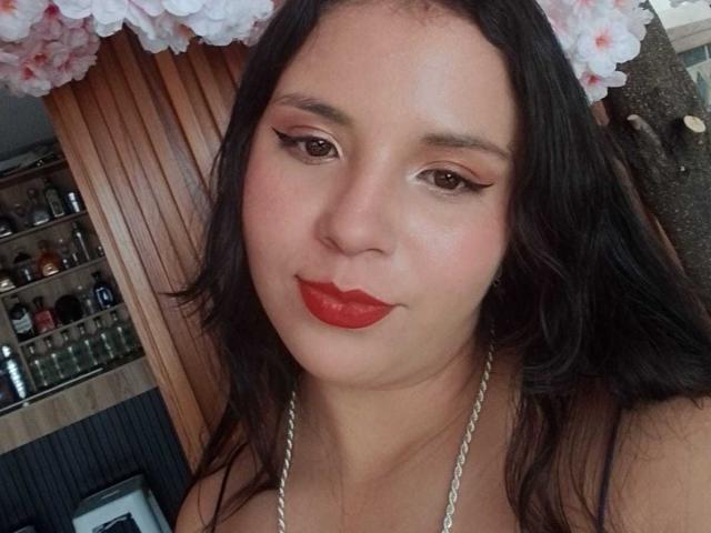 RosieTurner has a medium skin tone, with long, wavy black hair. She has thick, well-groomed eyebrows, subtle eye makeup with winged eyeliner, and bright red lipstick. Her expression is neutral with a slight smile. She's wearing a silver chain necklace. The background includes a wooden shelf with various bottles and a floral decoration above her head. The lighting is soft, highlighting her facial features. The image has a casual, candid style.