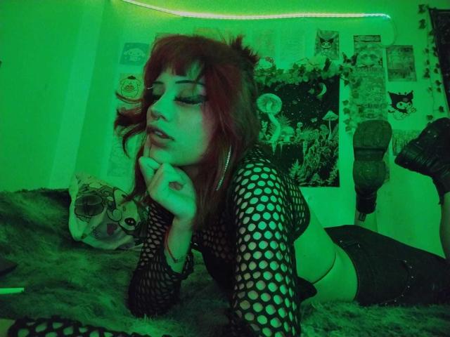 DollyBones with red hair, fishnet top, and midriff, lying on green-lit bed, hand on chin, looking seductive, green glow, furry blanket, and skull pillow in background.