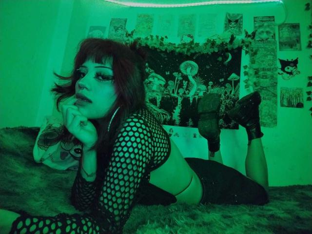 DollyBones with dark red hair, wearing a black fishnet top, lying on a furry rug, her hand near her mouth, green-tinted, showing her tits and ass.