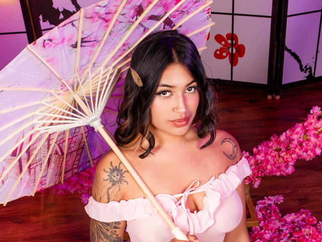 KatianPierce with dark hair, pink off-shoulder top, holding pink umbrella, tattoos on right shoulder, pink flower petals, wooden floor, purple and pink lighting, looking at camera, smiling.