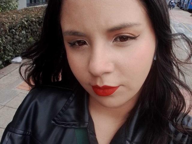 RosieTurner has medium skin, black wavy hair, and red lipstick. She's wearing a black leather jacket with green straps, and her eyelashes are prominent. RosieTurner looks down.