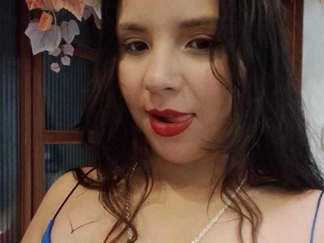 RosieTurner has medium brown skin, long wavy black hair, and red lipstick. She's wearing a blue spaghetti strap top and a silver necklace. Her eyes are slightly downcast, and she's sticking out her tongue slightly. The background has a wooden door with floral decorations. The image is a close-up, focusing on RosieTurner's upper body and face. She looks relaxed and playful. The photograph uses soft lighting to highlight her facial features.