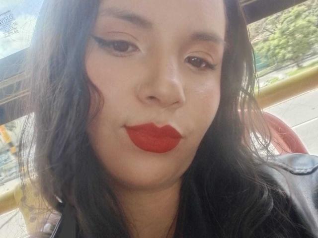 RosieTurner with red lipstick, black eyeliner, and black wavy hair, slightly pouting her full ass-shaped lips, wearing a black jacket, in a sunny outdoor setting.