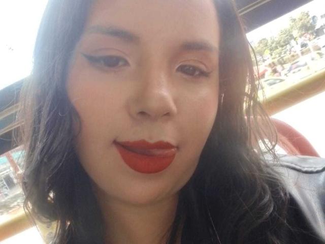 Close-up selfie of RosieTurner with medium brown skin, dark wavy hair, and red lipstick. She has black eyeliner with a sharp wing, slightly parted lips, and a subtle smile. She wears a black jacket. The background is blurred, showing a bright outdoor scene with greenery and a building. Her face is centered, and the lighting is natural. RosieTurner's makeup and lipstick are the most prominent features.