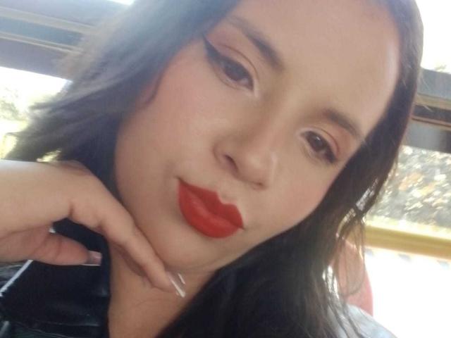 RosieTurner with red lipstick, black hair, and long eyeliner, resting her chin on her hand, showing off her shiny nails, wearing a black top, looking at the camera.