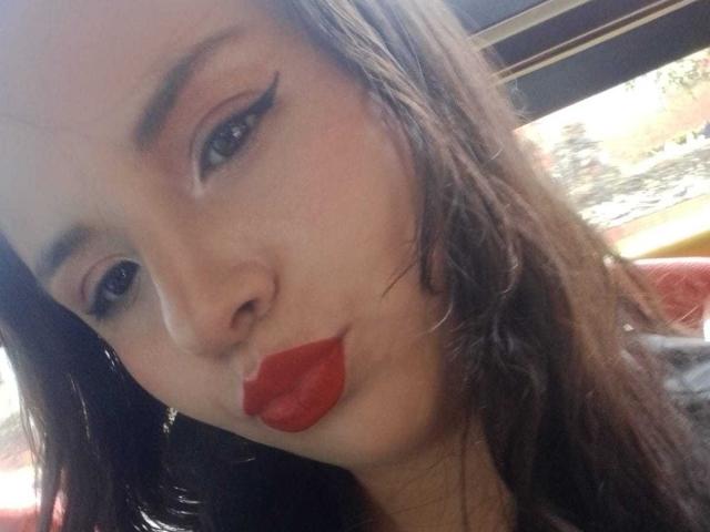 RosieTurner with red lipstick, black eyeliner, and brown hair, pouting her full, glossy, red lips. Her tits are slightly visible under a red top. She's looking at the camera.