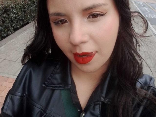RosieTurner with red lipstick, black eyeliner, and black leather jacket, tongue slightly out, long wavy black hair, green strap visible, looking at camera with a slight smirk.