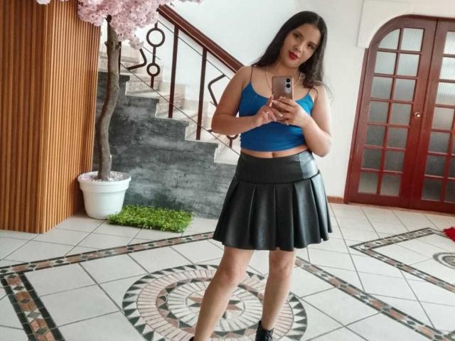 RosieTurner with dark hair, blue top, and black leather skirt, holding phone, standing in tiled room, pink cherry tree, wooden wall, red doors, and staircase in background.