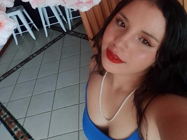 RosieTurner, a young Asian woman with long black hair and fair skin, takes a selfie. She wears a blue strapless top and a silver necklace, with bold red lipstick. Her brown eyes look slightly upwards. The image has a casual, slightly angled perspective. The tiled floor and white barstools in the background are partially visible. Pink flowers add a soft touch to the upper left. The overall composition is simple and focused on RosieTurner's face and upper body.