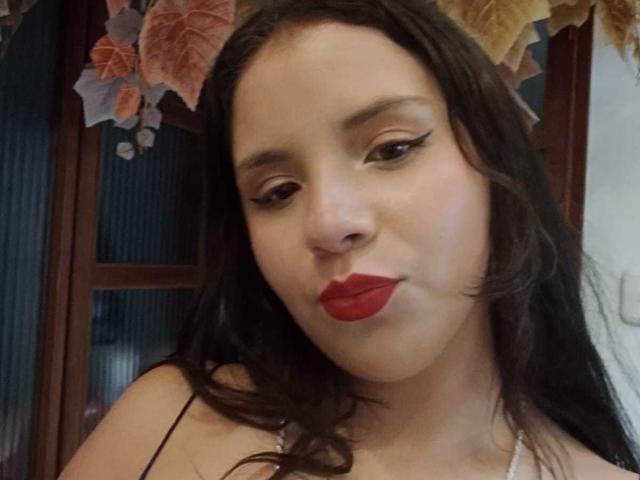 RosieTurner has red lips, black eyeliner, and long wavy dark hair, looking slightly to the side with a subtle pout. Her boobs are partially visible in a black top.