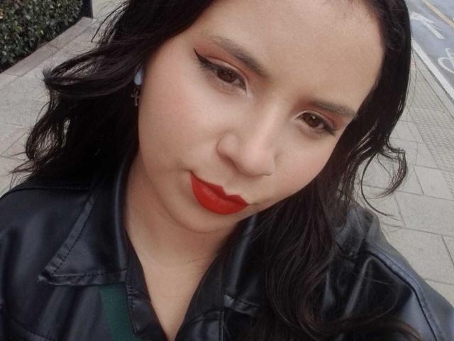RosieTurner has red lipstick, black eyeliner, and wavy black hair. She's wearing a black leather jacket with green straps. Her ass is not visible.