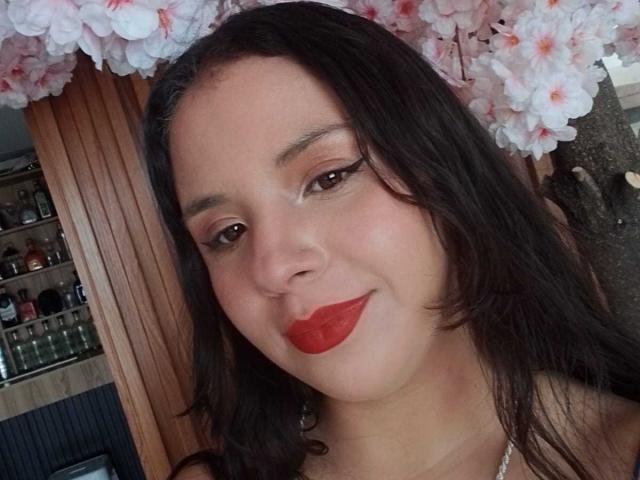 RosieTurner with red lipstick, dark wavy hair, and pink cherry blossom headpiece, smiling slightly. She has brown eyes, subtle makeup, and a small nose. Her tits are not visible.