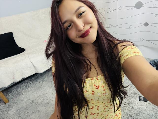 MarianaVargasX with long dark hair, red lipstick, nose ring, and floral dress, smiling at the camera, showing her tits in a selfie.