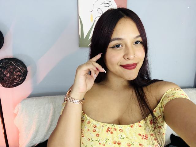 MarianaVargasX with medium brown skin, long black hair, red lipstick, floral off-shoulder top, smiling, touching her cheek, pink bracelet, cute, sexy, tits slightly visible, soft light.
