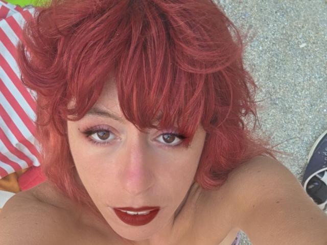 AnnarellaPikadZ con capelli rossi ricci, rossetto rosso scuro e trucco occhi rosa, che guarda verso l'alto con gli occhi socchiusi, indossa una maglietta colorata e giace su un asciugamano a righe..