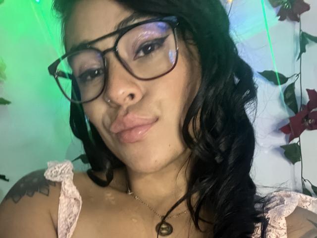 StellaBeckker with black glasses, pouty pink lips, and wavy black hair, wearing a lace top, showing tattoos on her shoulders, with a small nose ring.