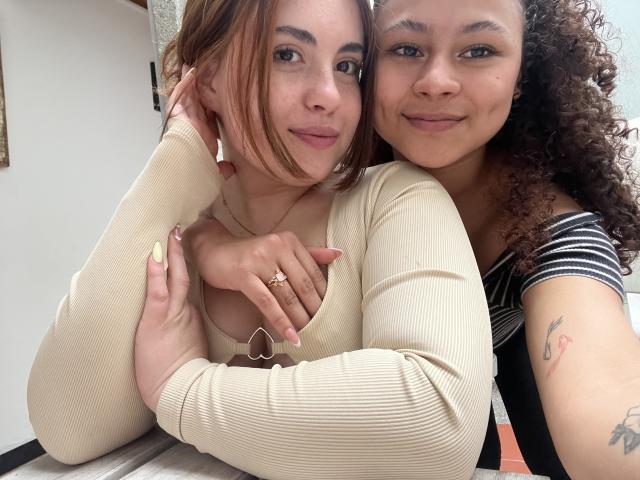 Two women in a close-up selfie. SofyViky on the left has light brown hair, wearing a beige ribbed long-sleeve shirt, holding a gold ring with a small heart, and a gold necklace. She has light skin and a slight smile. SofyViky on the right has curly dark brown hair, wearing a black and white striped shirt, with a tattoo on her right arm. Both women have their arms around each other, with SofyViky on the right resting her head on SofyViky's shoulder. Bright, natural light from the left.