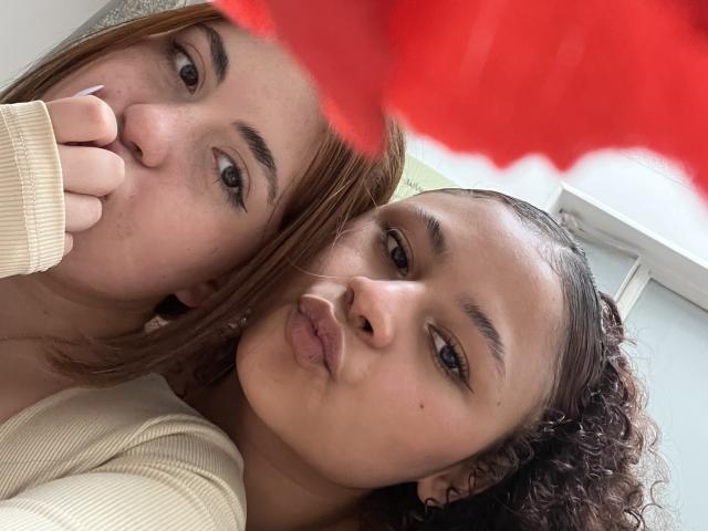 Close-up selfie of SofyViky. Two women with light brown skin and curly hair. One with straight, shoulder-length hair, the other with curly, chin-length hair. Both have dark eyebrows and are looking at the camera. The woman with straight hair has her finger near her mouth, while the curly-haired woman is puckering her lips. Both wear beige ribbed tops. Red, blurry flower in foreground. Soft natural light from the right. Simple, clean background.