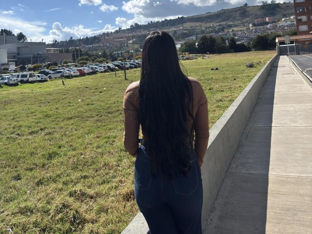 MiaDuboisX with long black wavy hair, tan top, and tight blue jeans, standing with her back to the camera, hands in pockets, on a concrete walkway, grassy field.