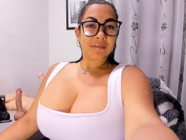 MellissaCortez with big tits, wearing glasses and a white tank top, has a cute smile, black hair in a ponytail, and a penis on the desk in the background.