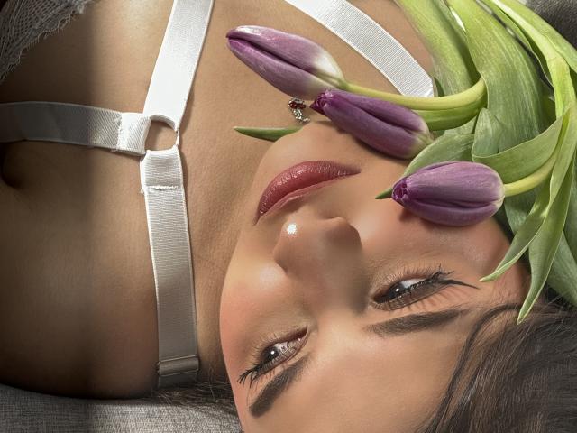 Close-up of MeganPaige's face with purple tulips on her forehead. She has dark eyebrows, long eyelashes, and glossy red lips. Her white strap harness is visible, with a silver ring in the center. Her skin is smooth and tan. The tulips are fresh, with green stems and purple petals. The composition is intimate, focusing on her facial expression and the tulips. The lighting is soft, highlighting her features and the petals.