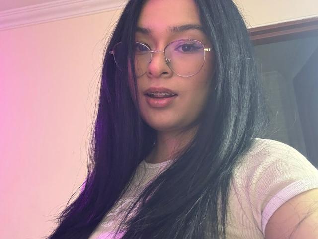 IsaDoux has long, straight black hair, wearing thin, round glasses with a gold frame. She has a light brown skin tone and is looking at the camera with a slight, confident smile. She's wearing a white, short-sleeved shirt. The soft pink and purple light casts a gentle glow on her face and hair. The image has a casual, selfie-style composition.