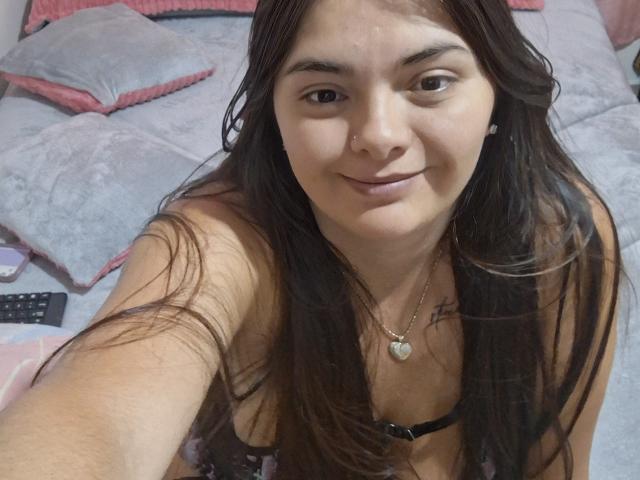 KatherinLatin with long brown hair, smiling, wearing a black bra, heart necklace, and nose ring, taking a selfie, showing her small tits and cute face.