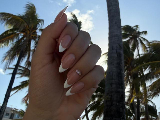 Hand shot from below, showing long, almond-shaped nails with white tips and a small, shiny diamond ring on the pinky finger. Skin is light and smooth. Palm trees and a clear blue sky are in the background, with sunlight casting shadows. The hand is slightly raised, with fingers curled gently. The image has a tropical, sunny feel. The palm trees are tall and slender, with green fronds. The composition is balanced, with the hand centered and palm trees in the background.