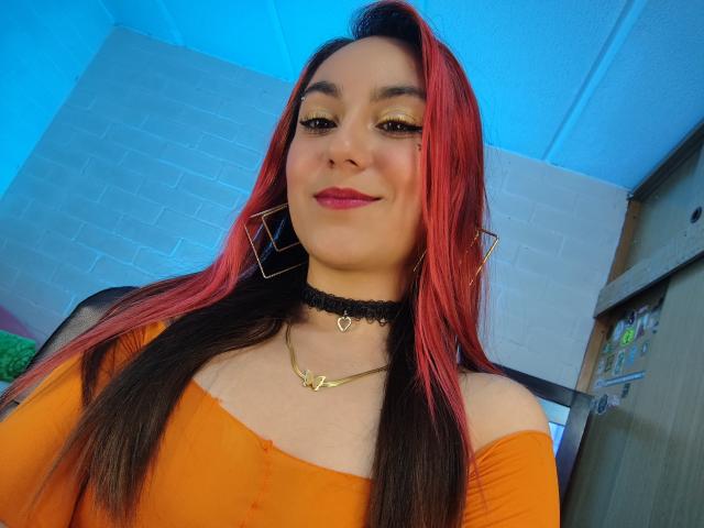 AmyStreetwise69 with red and black hair, orange off-shoulder top, black choker, gold necklace, and pink lipstick, smiling at the camera, wearing geometric earrings.