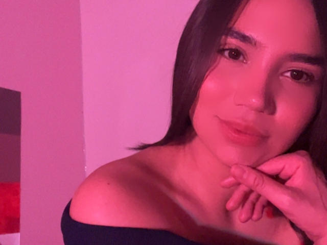 HannaCarlton with pink-tinged skin, brown eyes, and straight black hair, wearing an off-shoulder black top, gently resting her chin on her hand, smiling softly.