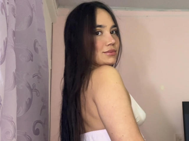 HannaCarlton with long black hair, white strapless top, looking over her shoulder, showing her back and a hint of her tits, in a pink-walled room, smiling softly.