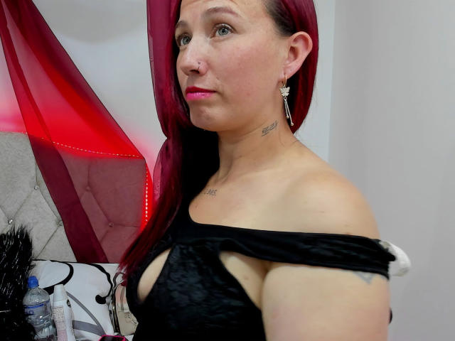 ChloeZenit with red hair, wearing a black, cut-out top, exposing her tits. She has pink lipstick, earrings, and a tattoo on her neck. Water bottle on desk.